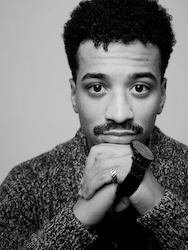A young Black man with a mustache poses in a black and white photo, resting his chin on his folded hands and wearing a textured sweater and a watch.