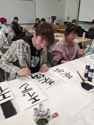 A young Caucasian male and an Asian female are intently practicing calligraphy in a relaxed and collaborative classroom atmosphere