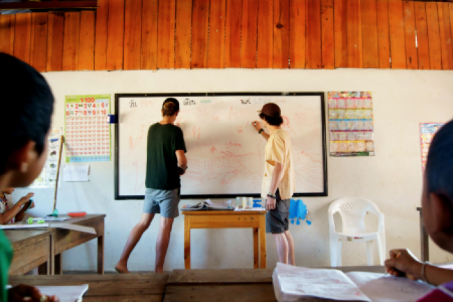 Two students in Thailand at a local elementary school at the whiteboard as local students look on.